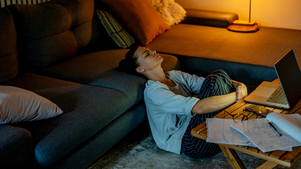 Junge Frau, die auf dem Boden vor dem Sofa sitzt, abends. Sie sieht müde aus.