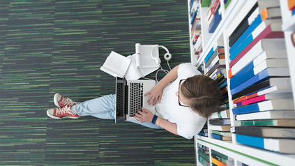 Junge Frau, die auf dem Boden der Unibiliothek mit ihrem Laptop und Kopfhörern an eine Regalwand gestützt sitzt und arbeitet.