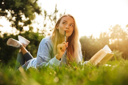Junge Frau liegt auf der Wiese mit Buch und Stift in der Hand und schaut verträumt auf