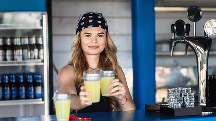 Junge Frau mit Bandana hinter Theke eines Getränkestandes auf Festivalgelände.