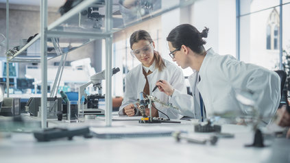 Eine junge Frau und ein junger Mann, die nebeneinander in Laborkleidung in einem Labor arbeiten.