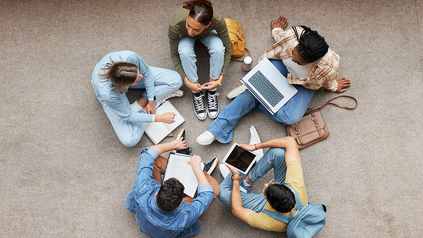 Fünf Studierende, die im Kreis auf dem Boden sitzen mit Laptops und zusammen arbeiten/diskutieren.