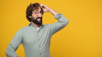 Nachdenklicher Mann mit Bart und Brille, der sich den Kopf kratzt, vor gelbem Hintergrund.