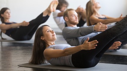 Fünf junge Menschen machen eine Pilates-Übung am Boden liegend.