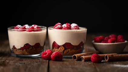 Zwei Gläschen gefüllt mit Spekulatius, Tiramisu und Himbeeren auf einem Holztisch drapiert.