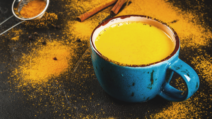 Tasse mit goldener Milch auf schwarzer Arbeitsplatte mit Kurkumaspritzern und Zimtstangen.