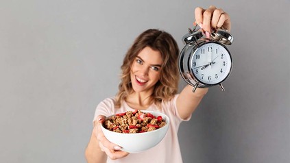 Junge Frau mit Müslischale in der einen und einer Uhr in der anderen Hand.
