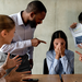Junge Frau in Büro, die von mehreren Arbeitskollegen angegangen wird.