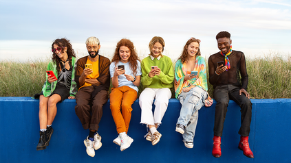 Gruppe diverser hipper junger Leute, die mit ihren Smartphones nebeneinander auf einer Mauer sitzen.