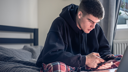Junger Mann, der im Schneidersitz in seinem Bett sitzt, Smartphone in der Hand, Laptop vor sich.