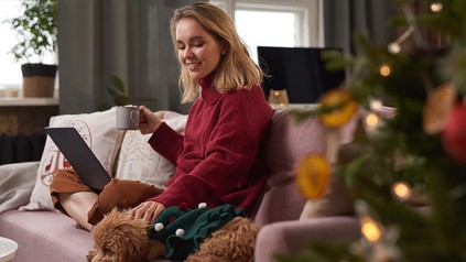 Junge Frau sitzt mit Hund auf dem Sofa und hat ihren Laptop auf dem Schoß.