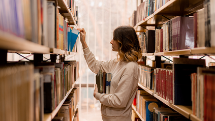 Junge Frau in Bibliothek mit Büchern in der einen Hand, während sie mit der anderen ein weiteres Buch aus dem Regel nimmt.