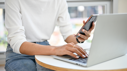 Ausschnitt von Torso einer jungen Frau, die mit einem Smartphone vor ihrem Laptop sitzt.
