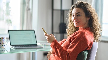 Junge Frau sitzt vor einem gläsernen Schreibtisch, auf dem ihr Laptop steht. Sie hat ihr Handy in der Hand und lächelt schräg nach vorne.