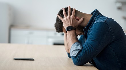 Junger Mann sitzt am Tisch, stützt seinen Kopf auf die Hände und sieht verzweifelt aus