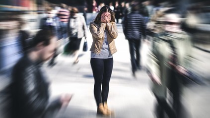 Junge Frau steht in einer Menschenmenge, hält sich die Augen zu. Die Menschen um sie herum sind verschwommen.