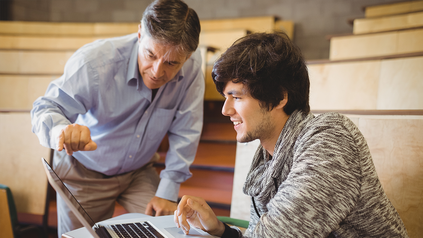 Dozent mit Studierendem am Laptop im Vorlesungssaal - sie sehen sich gemeinsam etwas an.