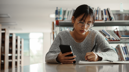 Studentin, die in der Bibliothek an einem Schreibtisch sitzt. Sie hält ihr Smartphone in der Hand und macht sich Notizen.