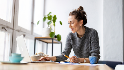 Junge Frau daheim an ihrem Schreibtisch mit Laptop vor sich.