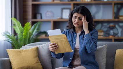 junge, indische Frau sitzt auf dem Sofa. Sie liest einen Brief und fasst sich schockiert an den Kopf.