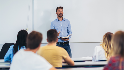 Junger Dozent im Hemd vor Studierenden. Er hält ein Buch in der Hand.