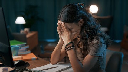 Gestresste Studentin sitzt am Schreibtisch vor Lernunterlagen. Sie hält sich die Hände an den Kopf uns ist verzweifelt.