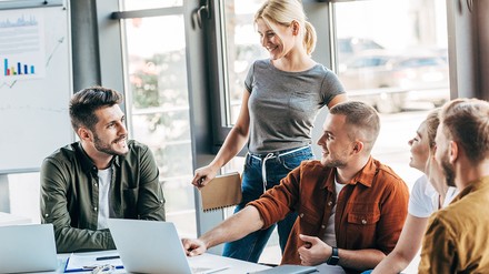 Fünf Studierende besprechen sich, auf dem Tisch stehen technische Geräte und Notizen, im Hintergrund ist ein Whiteboard mit Grafiken