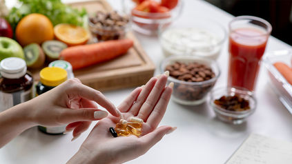 Frauenhände mit Pillen in der Handfläche, im Hintergrund Pillendosen, Obst, Gemüse und Nüsse.