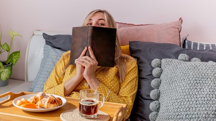 Junge Frau im Schlafanzug sitzt im Bett, vor sich Frühstück. In der Hand hält sie ein Buch, hinter dem sie sich versteckt.