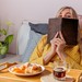 Junge Frau im Schlafanzug sitzt im Bett, vor sich Frühstück. In der Hand hält sie ein Buch, hinter dem sie sich versteckt.