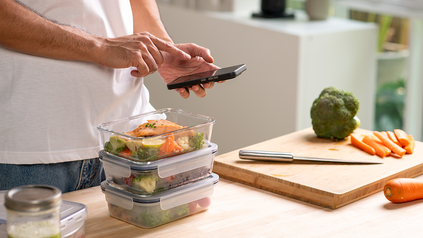 Junger Mann vor Kücheninsel, er packt gerade Essen in Tupperdosen und schaut aufs Handy.