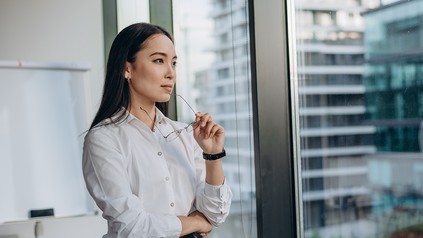 Junge Berufseinsteigerin steht nachdenklich am Fenster eines modernen Büros und blickt in die Ferne