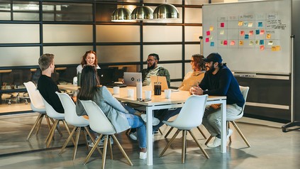 Junge Kolleg:innen sitzen an einem Tisch und arbeiten gemeinsam an einem Projekt, im Hintergrund ein Whiteboard mit vielen Post-its.