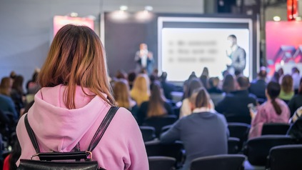 Ausschnitt von Vortrag aus dem Publikumsraum; vorne links junge Frau in rosafarbenem Hoody mit schwarzem Rucksack; der Kamera abgewandt.