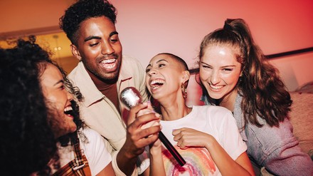Drei Junge Frauen und ein Mann halten gemeinsam ein Mirkofon, die Stimmung ist ausgelassen.