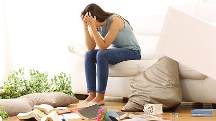 Juunge Frau sitzt verzweifelt in Ihrer Wohnung. Um sie herum liegen Bücher und Sofakissen verstreut.