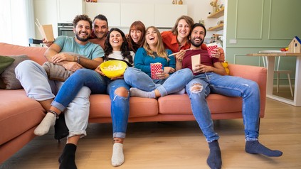 Eine Gruppe Studierender sitzt eng aneinander auf einem Sofa, das eigentlich zu klein für alle ist und schaut TV.