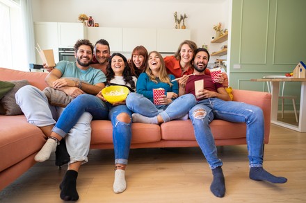 Eine Gruppe Studierender sitzt eng aneinander auf einem Sofa, das eigentlich zu klein für alle ist und schaut TV.