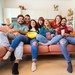 Eine Gruppe Studierender sitzt eng aneinander auf einem Sofa, das eigentlich zu klein für alle ist und schaut TV.