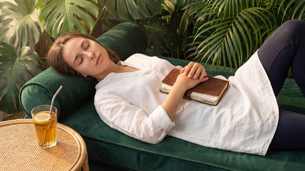 Junge Frau liegt auf einem grünen Sofa und schläft mit einem Tagebuch auf dem Bauch, neben ihr ein Tee.