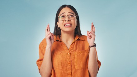 Junge Frau in gelben Shirt und Brille, schaut nach oben und hat die Finger gekreuzt.