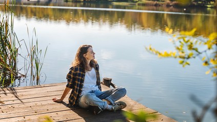 Junge Frau sitzt auf einem Steg und schaut entspannt auf das Wasser eines Flusses.