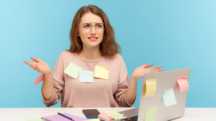 Junge Frau mit Brille sitzt ratlos am Schreibtisch vor ihrem Laptop, überall kleben bunte Post-it-Zettel.