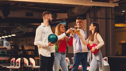 Freundesgruppe mit Bowling-Kugeln und Biergläsern in der Hand prostet sich auf Bowlingbahn zu.