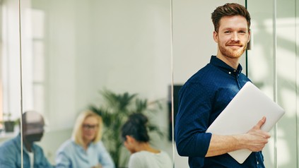 Junger Mann lächelt in die Kamera, er hält einen Laptop in der Hand. Im Hintergrund sieht man Menschen in einem beruflichen Setting.