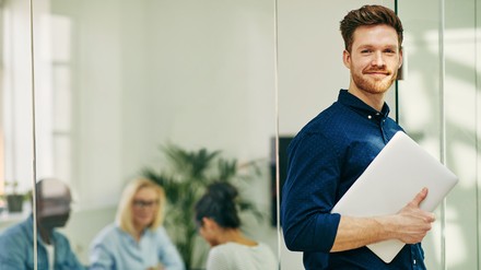 Junger Mann lächelt in die Kamera, er hält einen Laptop in der Hand. Im Hintergrund sieht man Menschen in einem beruflichen Setting.