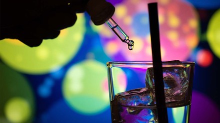 Glas mit Eiswürfeln und Pipette gefüllt mit Flüssigkeit, die von einer Hand darüber gehalten wird – vor buntem-Lichter-Disko-Hintergrund.