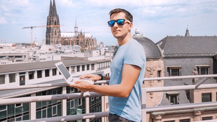 Ein junger Mann steht mit Laptop an einem Geländer und blickt auf Köln.