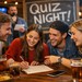 Eine Gruppe junger Erwachsener sitzt in einer Bar an einem Tisch und nimmt gemeinsam an einem Quizabend teil.