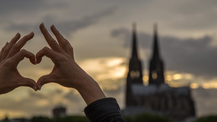Zwei Hände formen ein Herz. Im Hintergrund die Skyline von Köln.